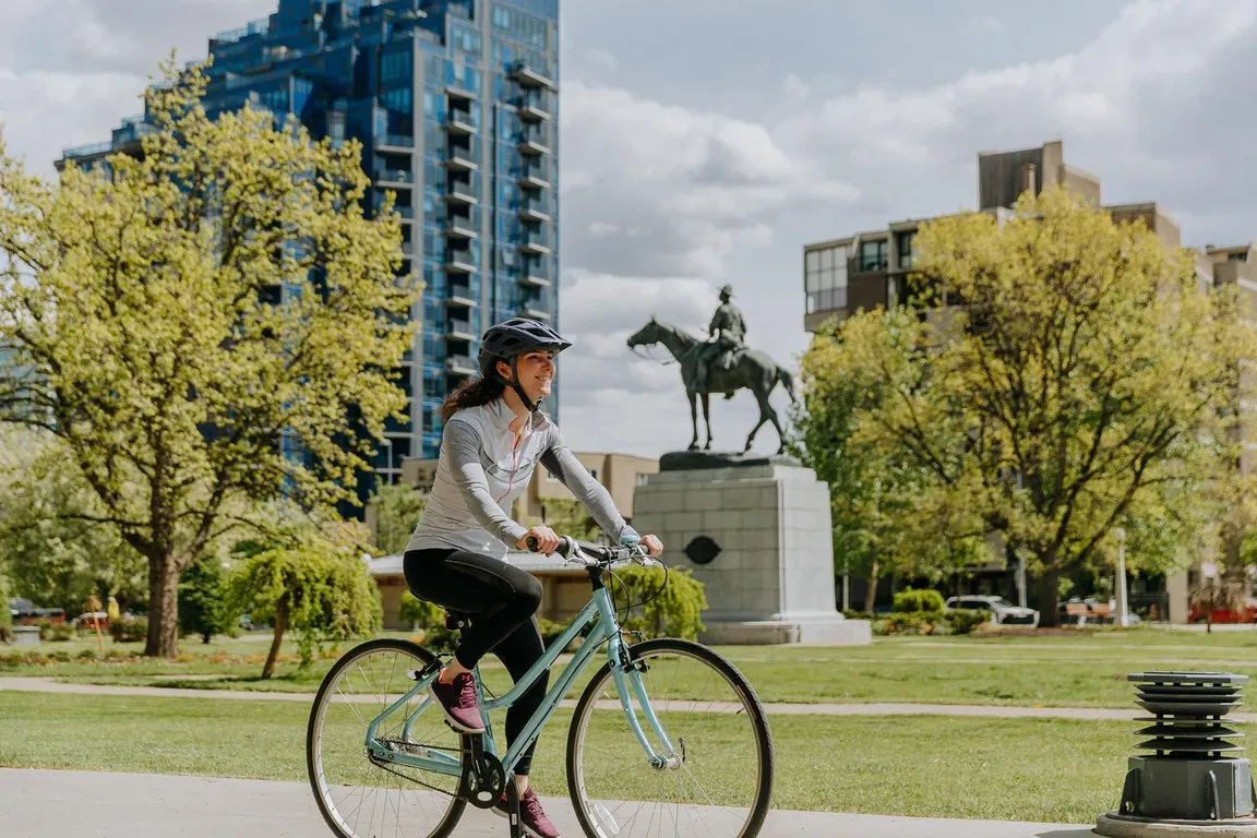 Woman riding a light blue bicycle past a horse statue in an urban park.
