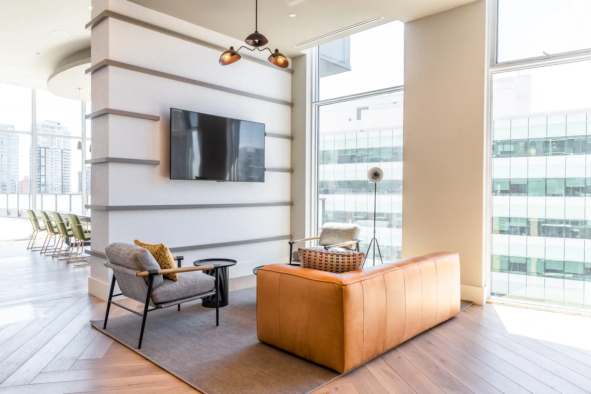 Modern lounge with brown sofa, gray armchair, wall-mounted TV, and large city windows.