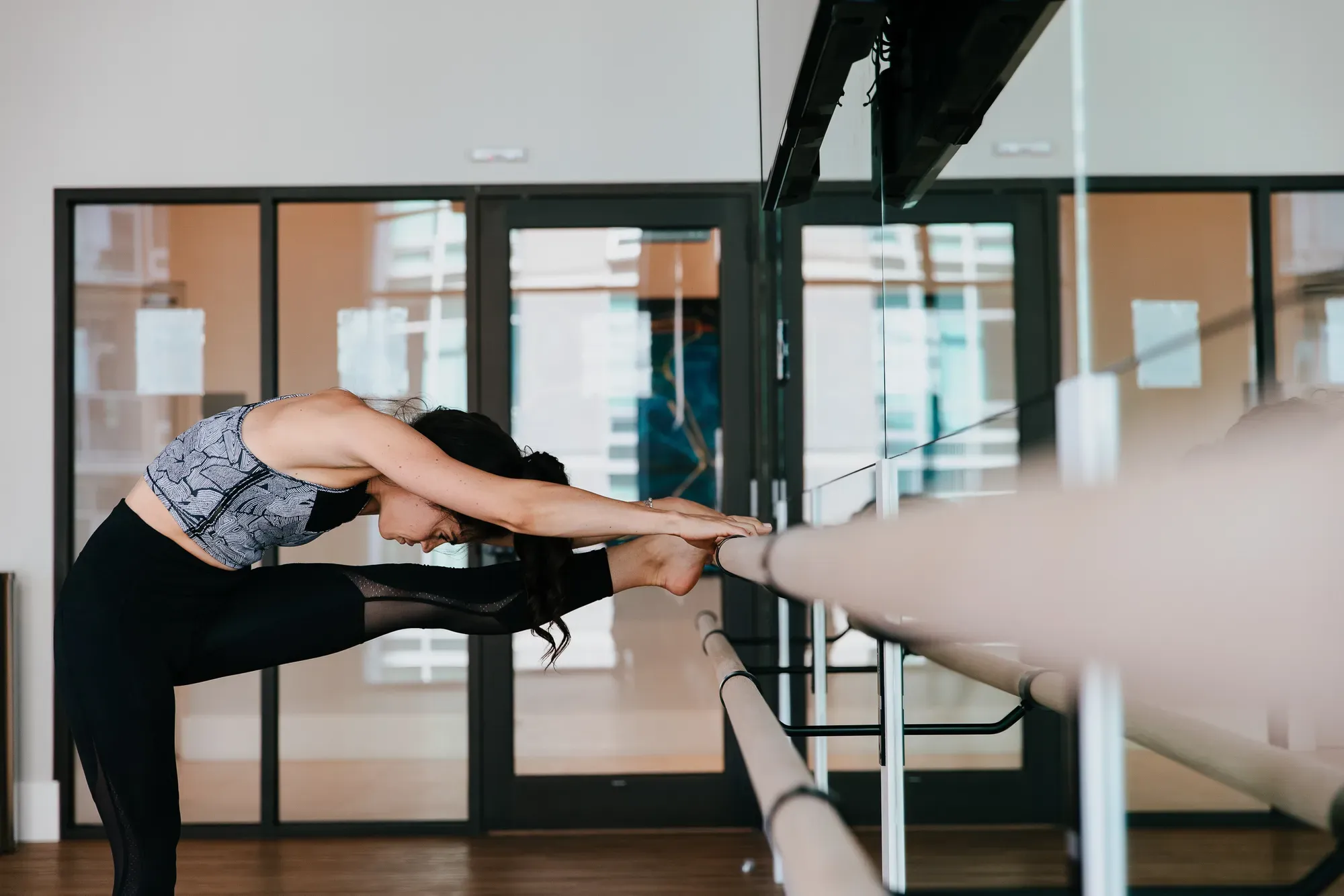 Woman in activewear stretches her leg towards a ballet barre in a studio.